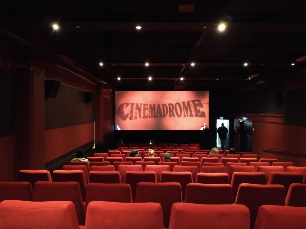 Kuva melko tyhjästä elokuvateatterista. Punaiset penkit, hämärä valaistus. Valkokankaalla elokuvafestivaali Cinemadromen logo.