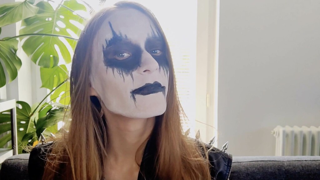 Black metal maskeissa oleva henkilö lähikuvassa ja katsoo hieman hymyillen kameraan